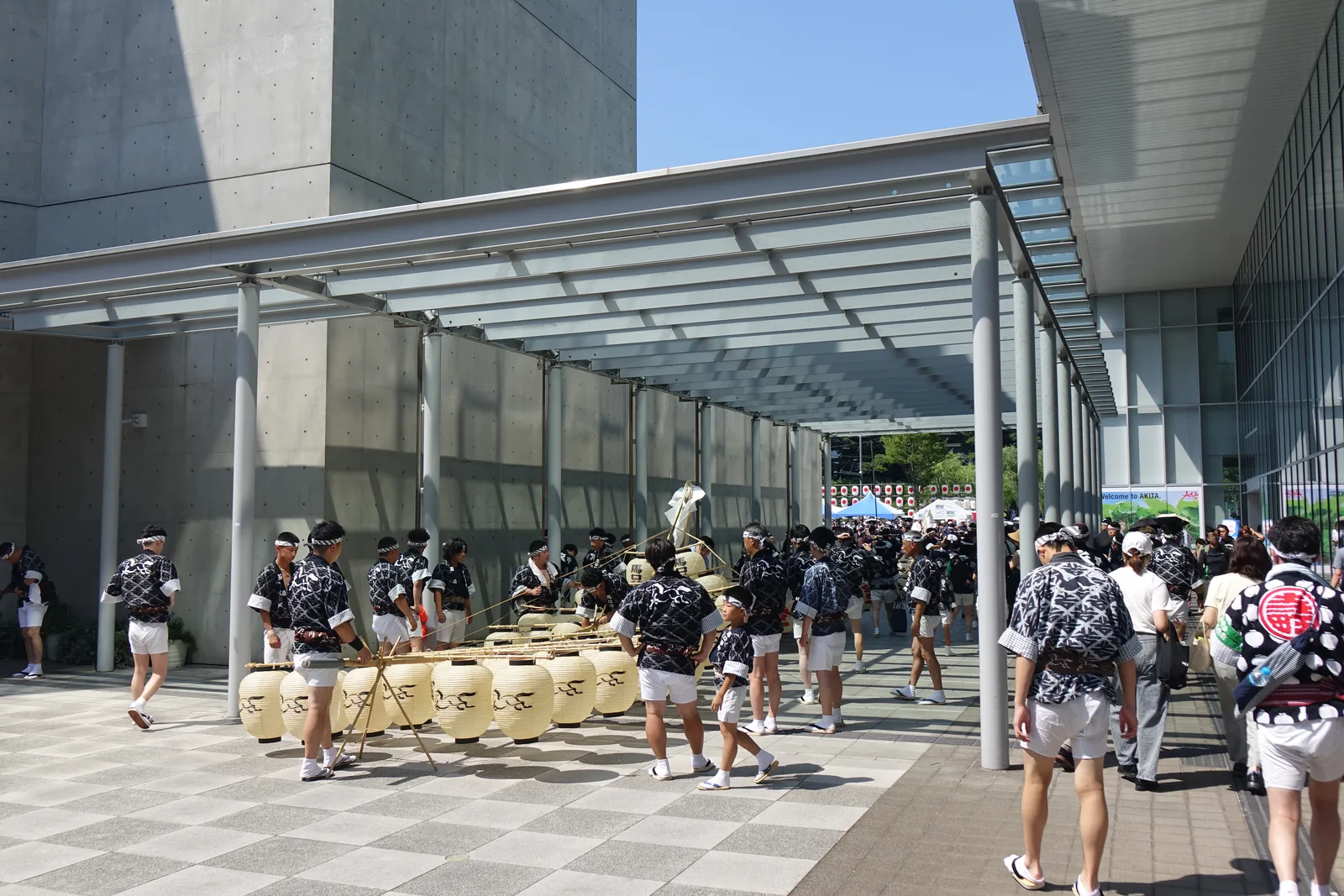 雨庇下的待命處，地面上堆放著竿燈的燈籠，身穿法被的隊員在陰影中走動，左側是美術館的清水混凝土牆