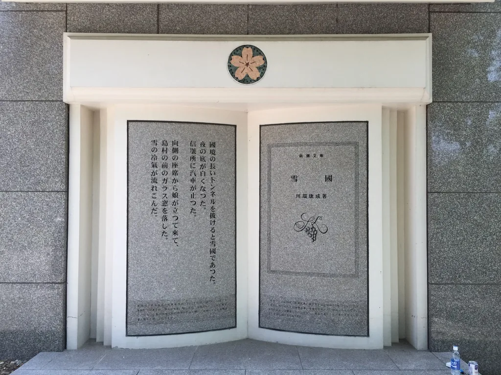 灰色石材外牆上嵌著翻開書本形狀的物件，白色的框中左右兩頁為石板，右頁刻著書名雪國與川端康成，左頁刻著小說的開頭，上方嵌著一枚櫻花紋章