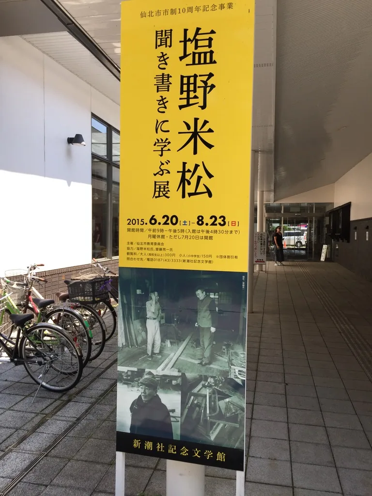 建築側面的步廊，上方是深出簷，地面鋪著石磚，左側停放著腳踏車，前景立著黃色的企畫展旗幟，盡頭是玻璃入口