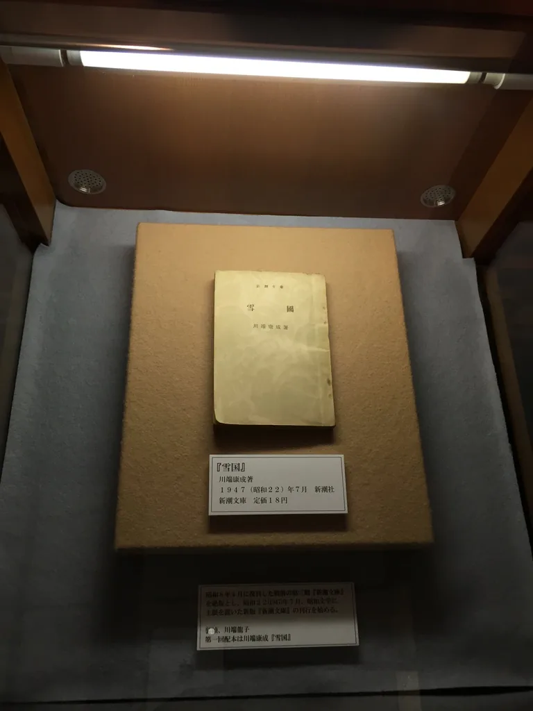 木框玻璃展示櫃中，淡褐色布面的台座上放著一本泛黃的文庫本，下方有說明牌，櫃頂設有燈具