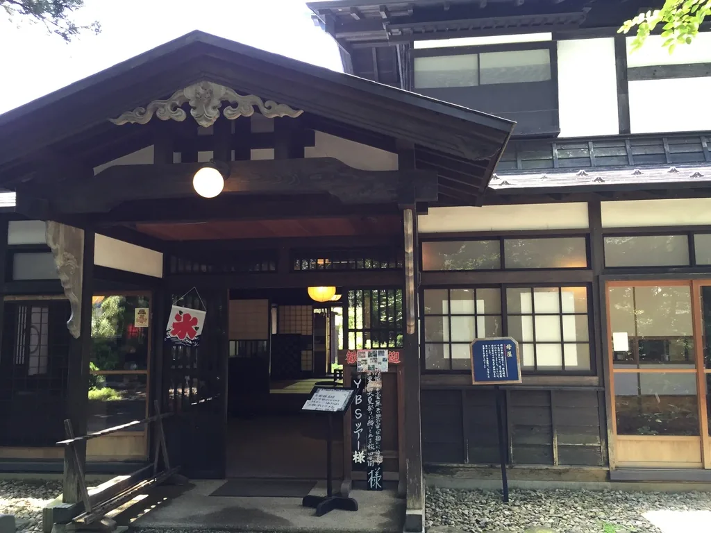 西宮家母屋的玄關，木造的山牆屋頂下方是寬大的入口，入口兩側為格子窗，門前立著告示牌與燈籠