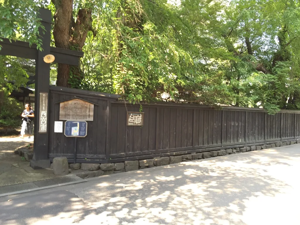 田町的道路旁，一道長長的黑色板牆沿著街道延伸，板牆之上是茂密的樹木，入口處立著木製的告示板
