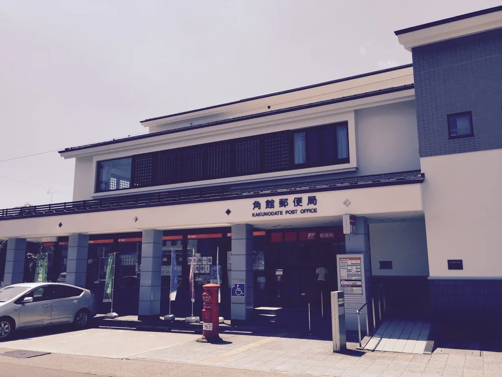 角館郵便局的正面，兩層的建築以白色牆面與深色的格子窗構成，一樓設有入口與紅色郵筒，前方是停車場