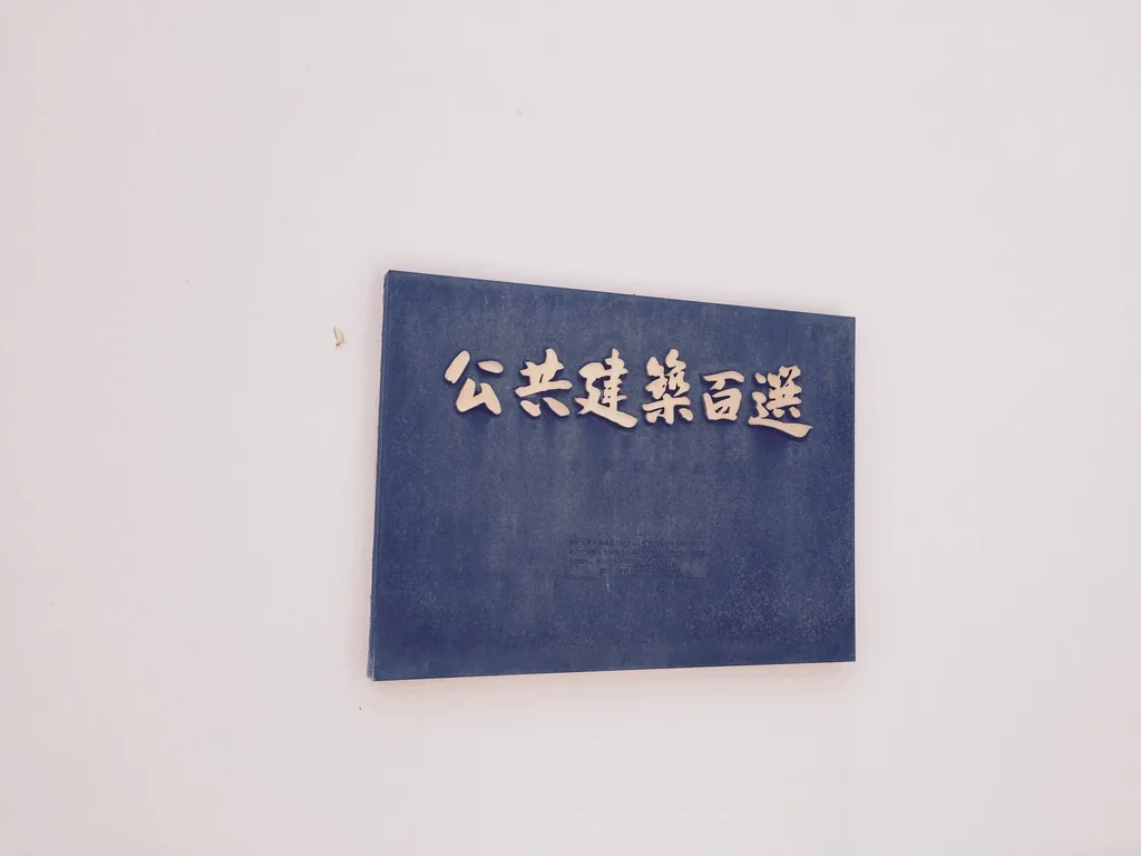 白色牆面上一塊深色的銘板，以金色字寫著公共建築百選