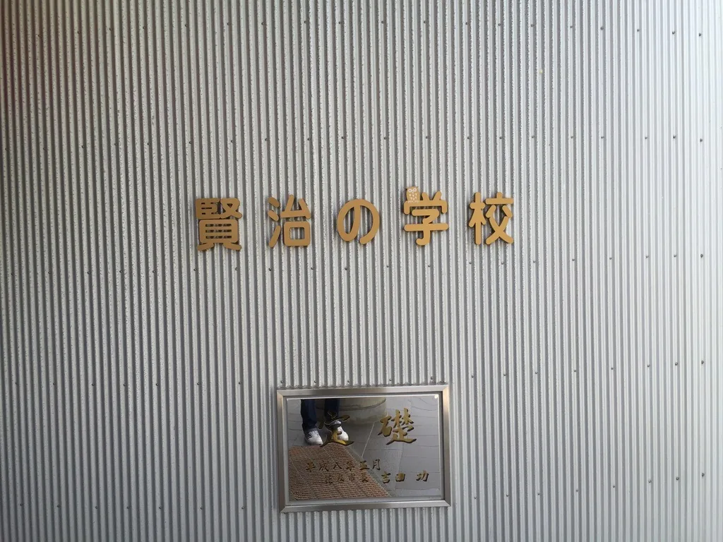 白色波浪板外牆上以金色字寫著賢治之學校，下方嵌著一塊不鏽鋼框的定礎板