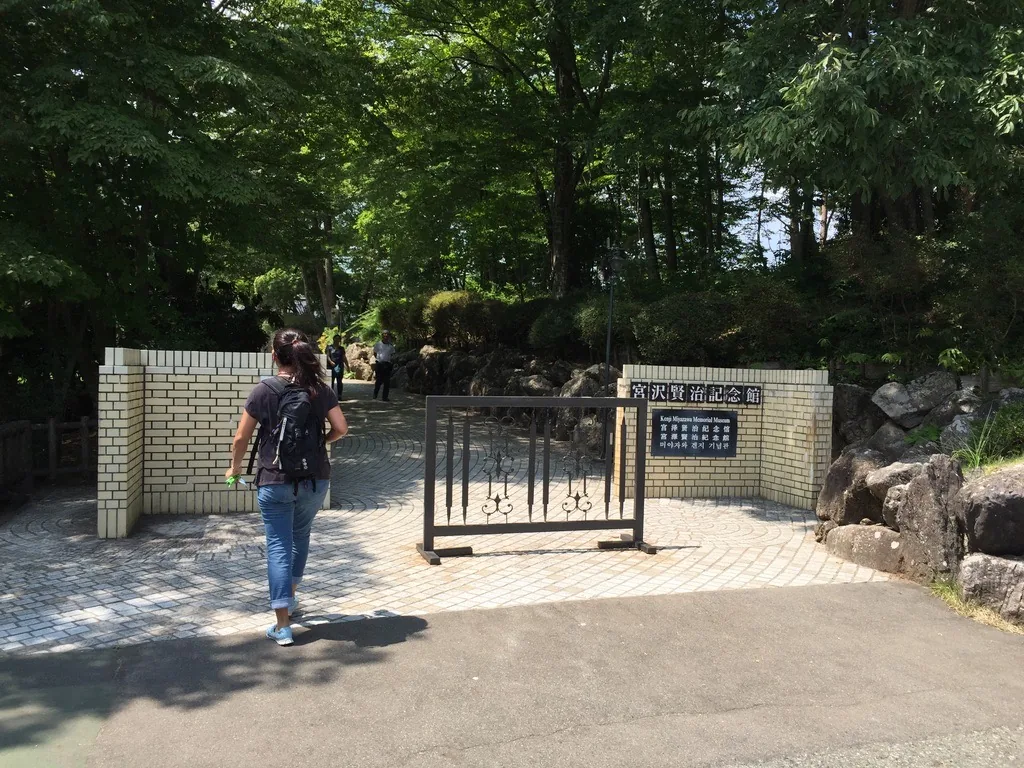 林間步道的起點，兩側立著低矮的淺黃色磚紋牆，右側牆上掛著館名銘板，步道向樹林中延伸
