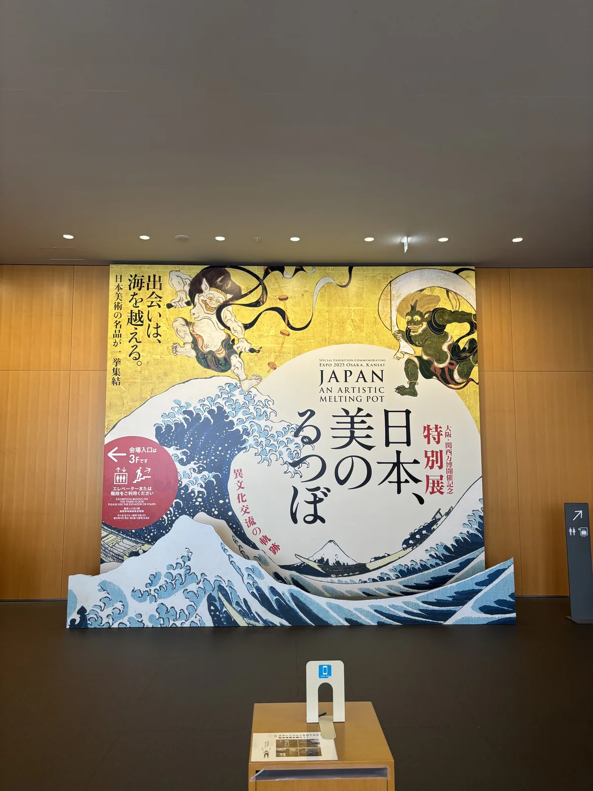 平成知新館一樓的展覽入口，一面高大的木質面板牆前，立著一幅以浮世繪巨浪與風神雷神為圖案的特別展大型看板，看板前方是一個小型的導覽架，天花排列著嵌燈