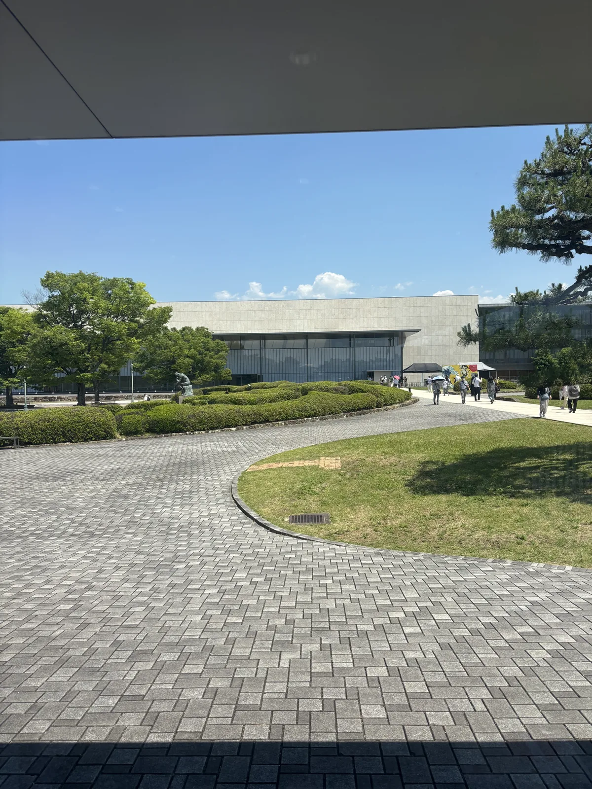 自南門入口望向園內，一條灰色石板鋪成的弧形步道穿過草坪與修剪成團的灌木，遠處是平成知新館的低矮量體與深出挑的屋頂，右側是一棵松樹的枝葉