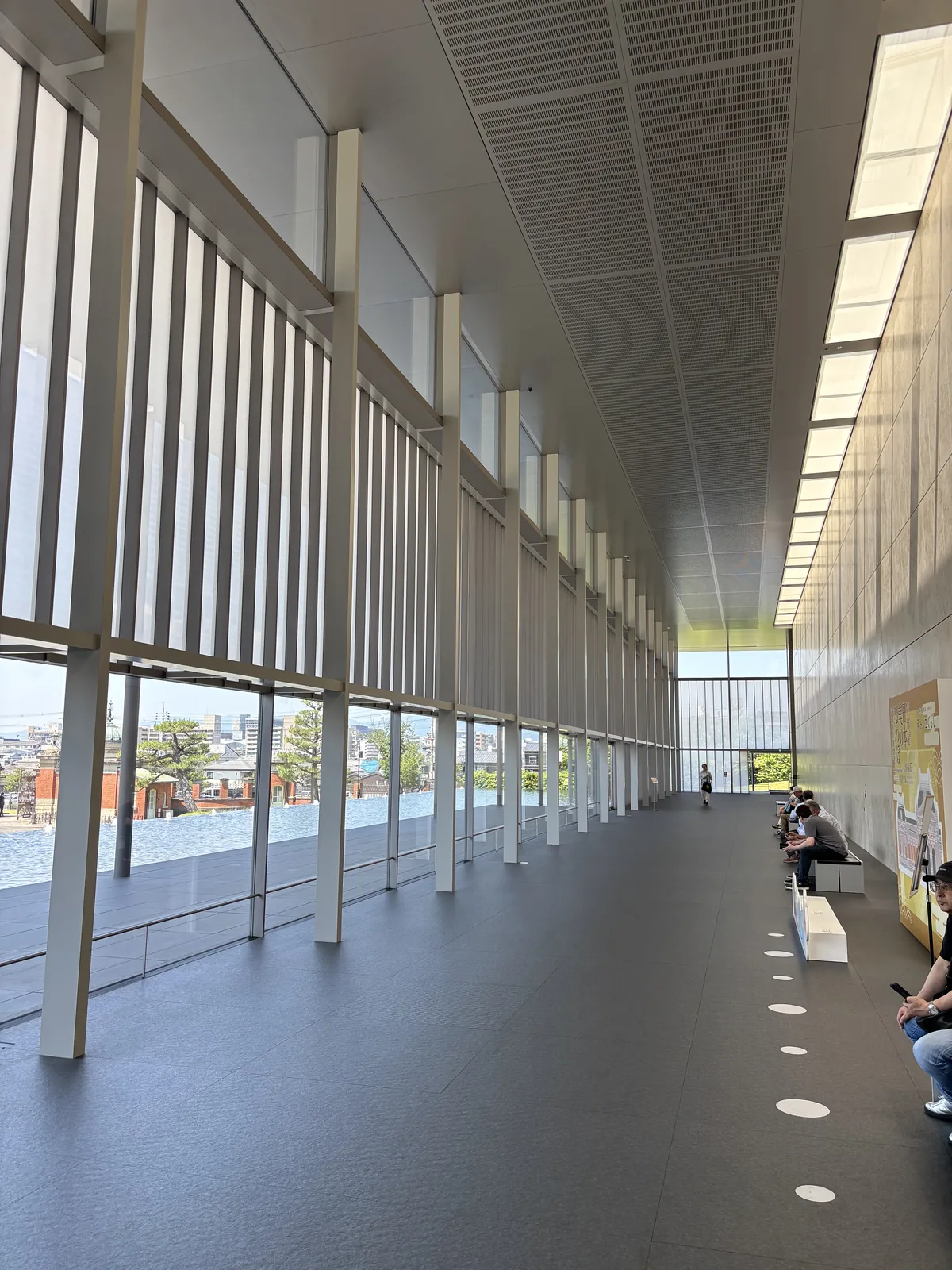 Grand Lobby 的長向內景，左側是一整列細長的直向 louver 與玻璃，louver 之外是水池與樹木，右側是淺色的石材牆面，天花是深色的有孔板並排列著一道長條的照明，遠處有幾位坐在長凳上的參觀者