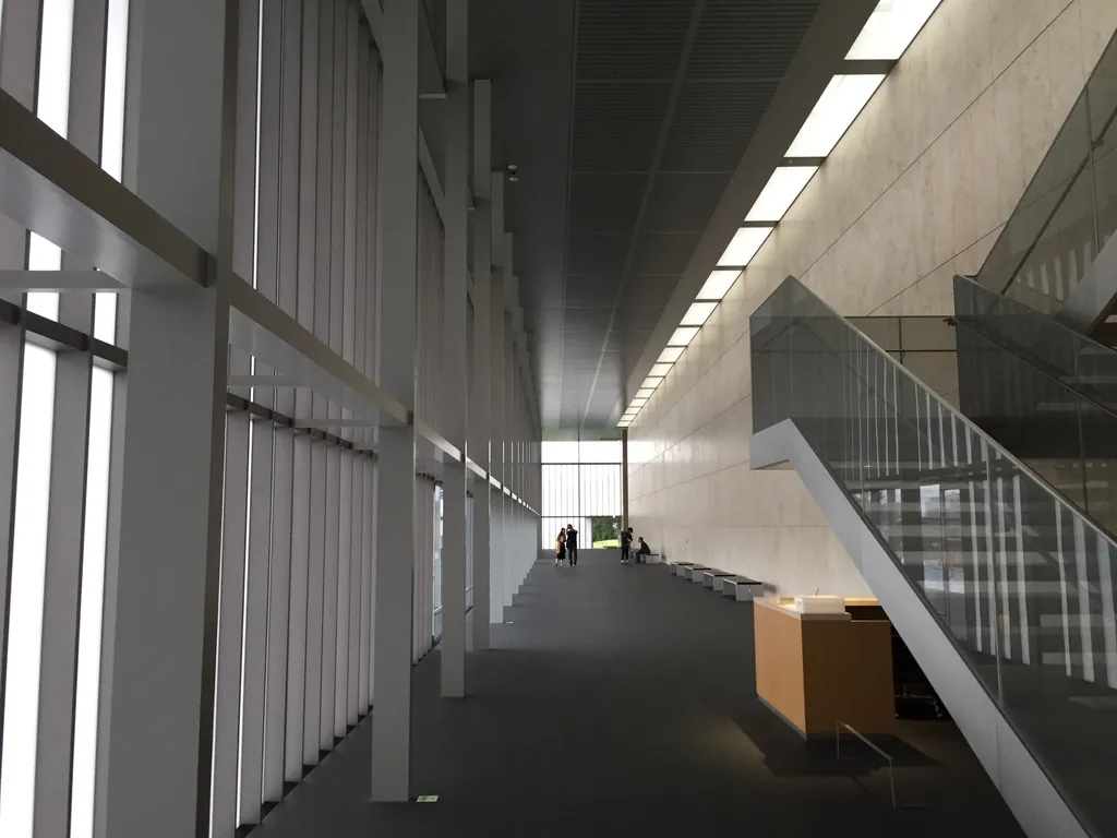 一樓 lobby 的內景，左側是一道深色的玻璃 louver 牆，右側是淺色石材牆與一道斜向上升的樓梯，樓梯旁是一個淺色木質的櫃台，lobby 的盡頭是一片明亮的玻璃開口，遠處有幾位參觀者