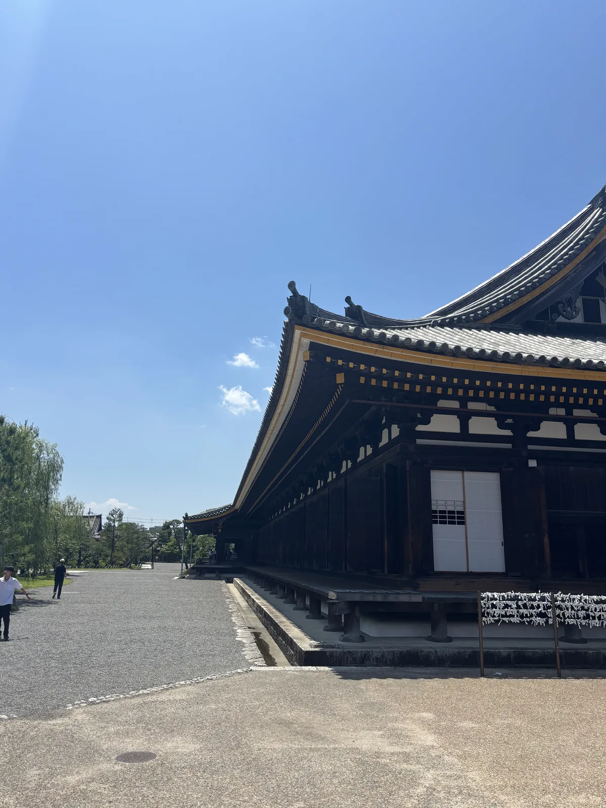 三十三間堂的本堂一角，一棟深色木造的細長堂宇向遠處延伸，屋簷深出挑，前方是礫石鋪成的廣場與幾位參觀者，天空晴朗