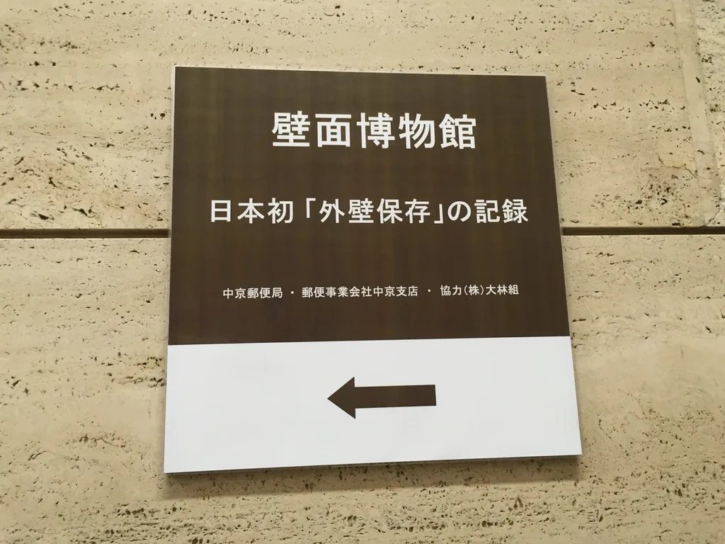 一塊褐色底的告示板，以白字寫著館名「壁面博物館」與副標「日本初外壁保存的記錄」，下方以小字列出中京郵便局、郵便事業株式會社中京支店與協力大林組，最下方是一個指向左方的箭頭，告示板貼在淺米色的石材牆上
