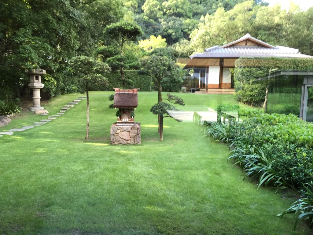 自地上棟的玻璃面望向庭園，前景是修剪過的灌木，中景是一片草坪、一座石燈籠與幾株修剪成層的松樹，草坪後方是一棟瓦葺屋頂的低矮和風建築，背景是濃密的山林