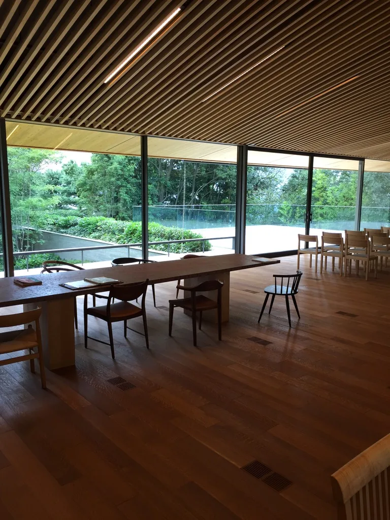 一樓 hall 的內景，天花是密排的細木條並順著屋頂向兩側傾斜，地面是深色的木地板，正面是整面落地玻璃，玻璃外是綠色的庭園，室內擺著一張長木桌與幾張木椅，遠處是成排的椅子