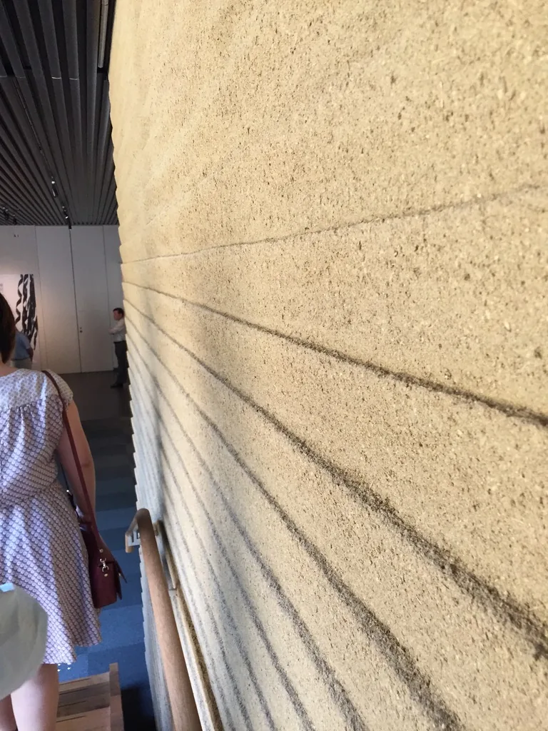 地下樓梯旁的土壁，一道粗糙的淺土色牆面占滿畫面右側，牆面上有幾道深色細線呈放射狀斜向延伸，左側是扶手、一位參觀者與遠處的展示空間，上方是深色的天花