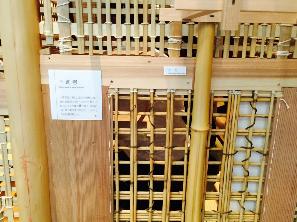 茶室 skeleton 模型的局部，竹子與細木條以繩紮成格狀的牆體骨架，中央是一扇下地窗的開口，左側一根竹柱與淺色木材的框架，上方貼著一張白色的說明牌