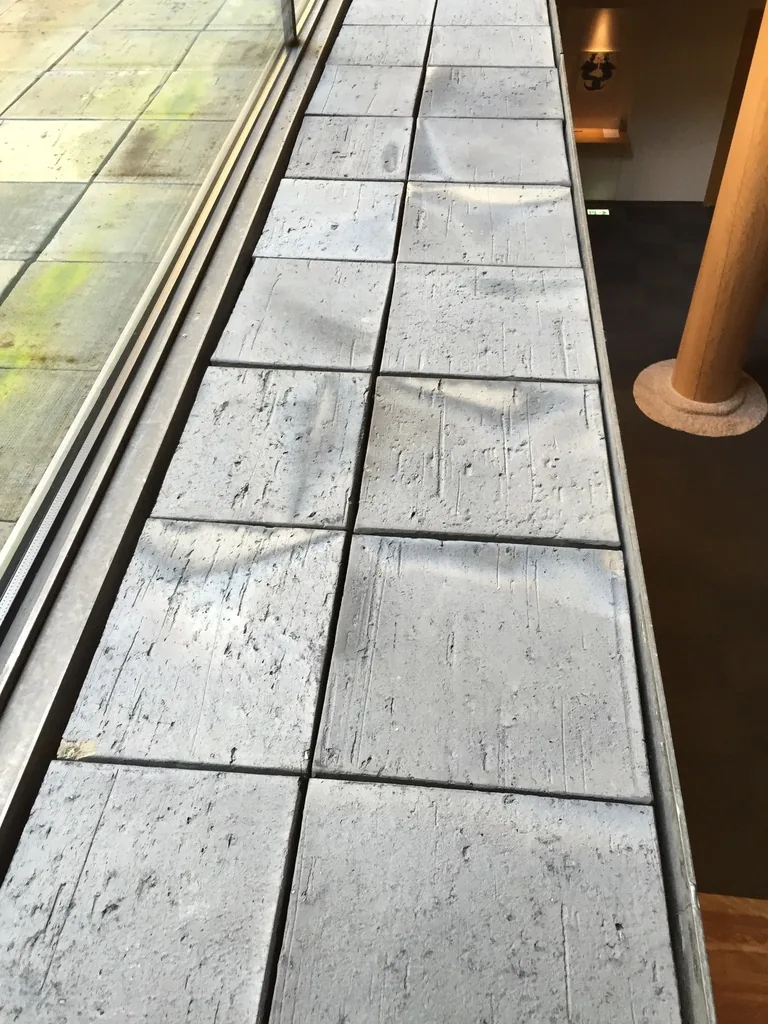 地下一層中庭沿著玻璃面的地面，灰色的方形瓦片鋪成兩列，瓦面帶著不規則的刷痕與斑點，左側是玻璃窗框，右側是室內的深色地面與一根木圓柱的底部