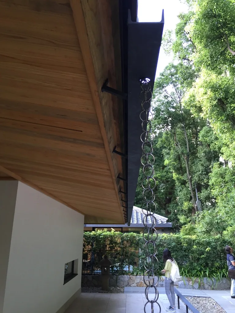 地上棟屋簷的端部，深出挑的木板天花自左上方伸出，簷端以深色金屬收邊，一條金屬鎖鏈自簷端垂下，右側是濃密的樹木，下方是白色的牆與綠籬