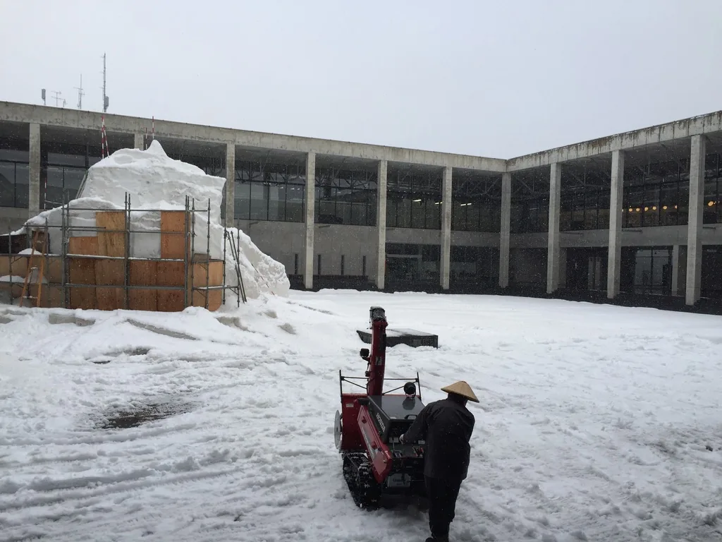 十日町 Kinare 的中庭，積雪上立著一座以鷹架與木板模框圍起的大雪塊，前景是一台紅色的履帶式除雪機與一位戴斗笠的居民，背景是環繞中庭的二層回廊，清水混凝土柱以等間距排列，屋頂是一道薄的水平線