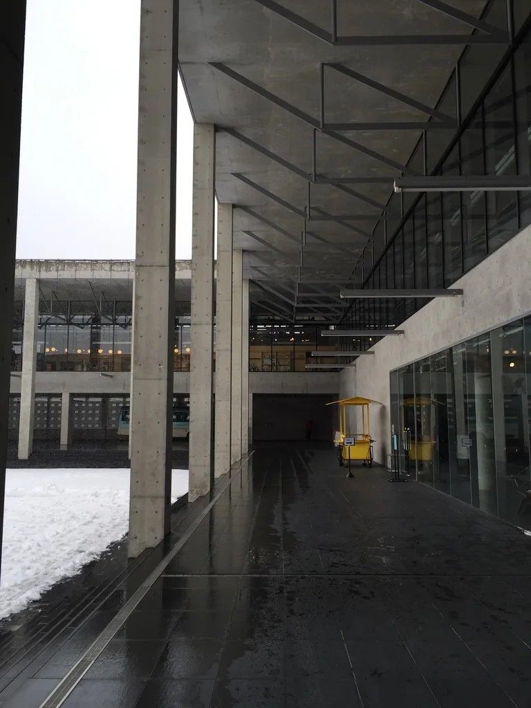 Kinare 一樓的回廊，左側是一排清水混凝土方柱，柱外是積雪的中庭，右側是玻璃的店面與一道混凝土牆，天花是平坦的板面，上面可見斜向交錯的線條，地面是濕亮的深色石材，遠處有一座黃色的小攤車