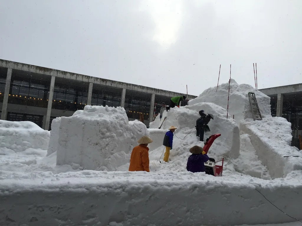 中庭裡的雪雕製作現場，一座以鷹架與木板圍起的大雪塊後方是二層的回廊，前景是一台紅色的履帶式除雪機，一位穿黑色外套、戴斗笠的人正在操作