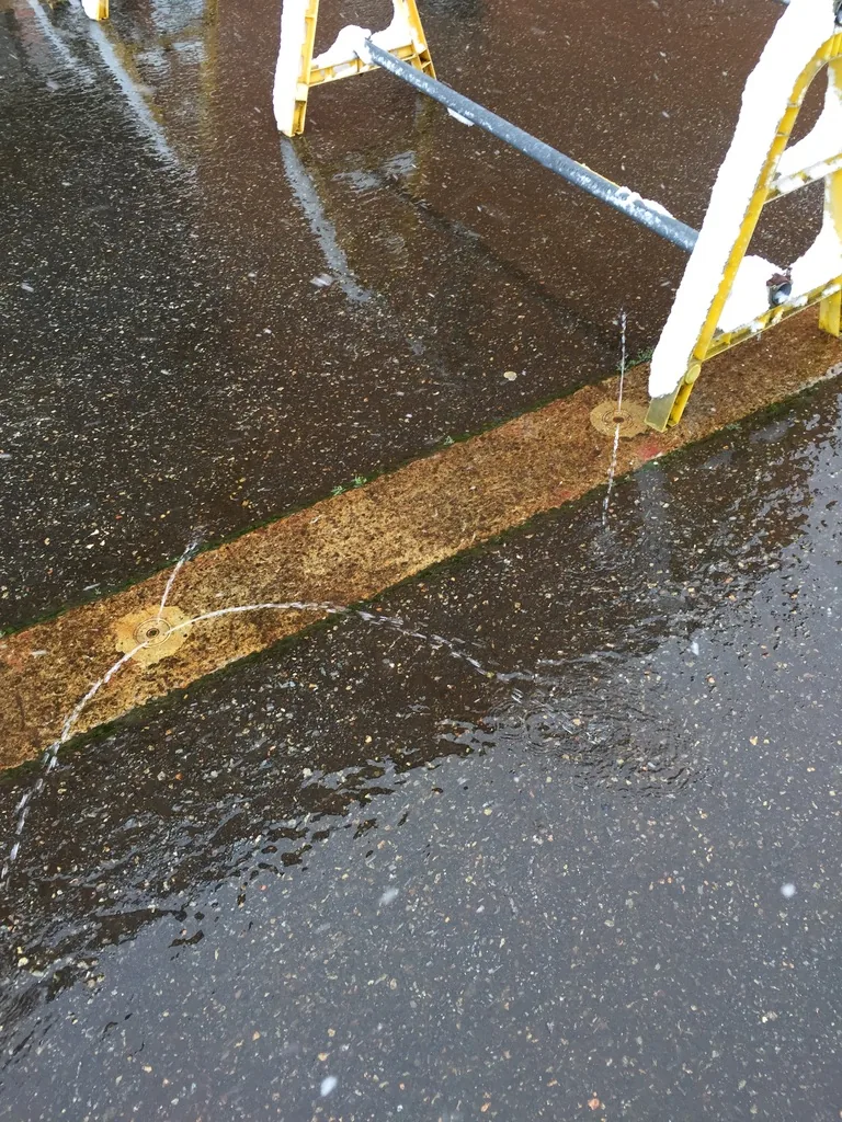 館外鋪面上的消雪設備，金屬噴嘴自地面向外噴水，水在深色的柏油上散開，旁邊是一架覆雪的黃色梯架