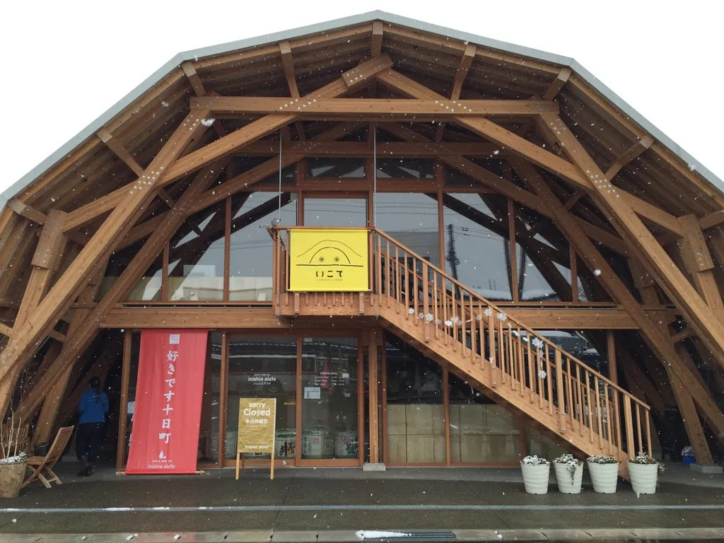 十日町產業文化發信館 Ikote 的正面，巨大的木造拱形構架從地面立起，正面中央是一座外露的木造樓梯通往二樓，樓梯上方掛著一面黃色的館名布幕，一樓是玻璃的店面，門前排著幾個白色的花盆，左側掛著一面紅色的直幅