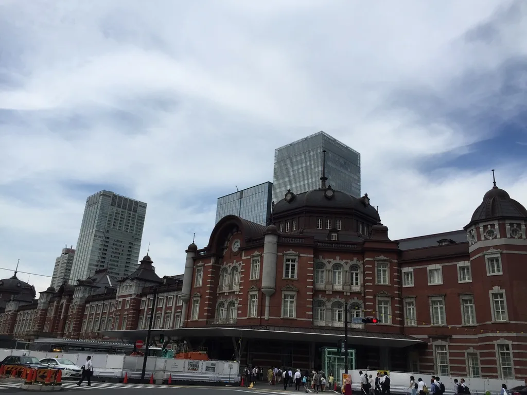 東京站丸之內站舍的正面，紅磚與白色石材構成的三層立面中央有一座塔屋，右端是圓頂，站舍後方是數棟玻璃高樓，前景是白色的施工圍籬與行人