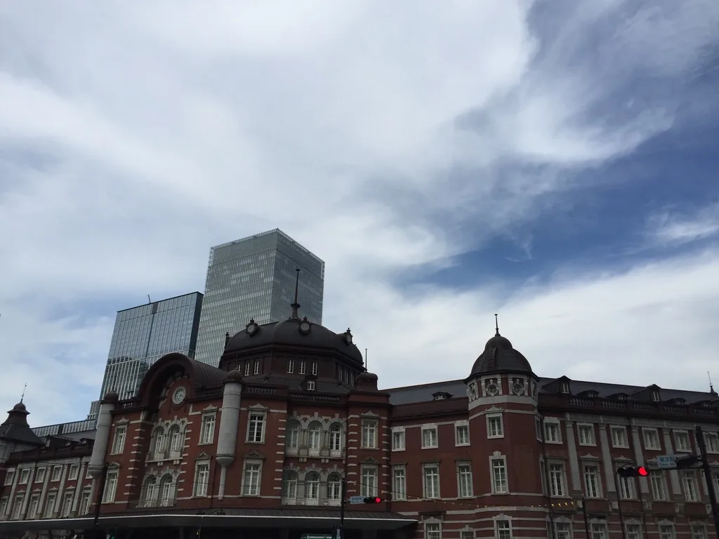 東京站丸之內站舍的中央與南段，紅磚立面上排列著白色石材收邊的窗，中央塔屋與南側圓頂並立，站舍後方是兩棟玻璃高樓，天空中有大片的雲