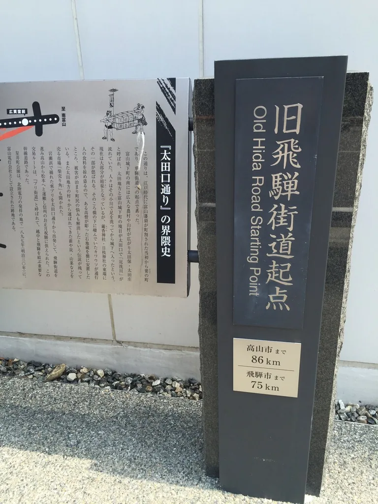 TOYAMA Kirari 外牆前的舊飛驒街道起點碑，深灰色的石柱上刻著館名與英文，下方的銘板寫著到高山市八十六公里、到飛驒市七十五公里，左側是一塊說明板，背景是白色的外牆面材