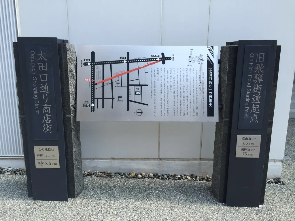 TOYAMA Kirari 外牆前的兩座石柱與一塊說明板，左柱寫著太田口通商店街與英文，下方銘板寫著此地海拔十一公尺、距海岸八點五公里，右柱是舊飛驒街道起點碑，中間的不鏽鋼說明板上畫著周邊的街道圖與太田口通的歷史