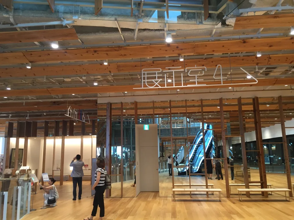 美術館區的展示室入口，天花上方懸著以白色細線寫成的大字「展示室4」與箭頭，下方是一排直立的杉木柱與一扇白色的門，右後方可見電扶梯，地面是木地板，幾位參觀者站在入口前