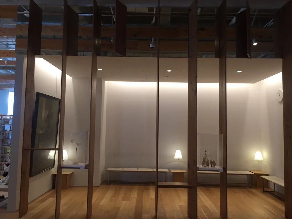 以玻璃隔開的展示室，前方是一排直立的杉木 louver，louver 之間可見展示室內白色的牆、間接照明的天花、幾件放在玻璃罩內的展品與一幅油畫，地面是木地板