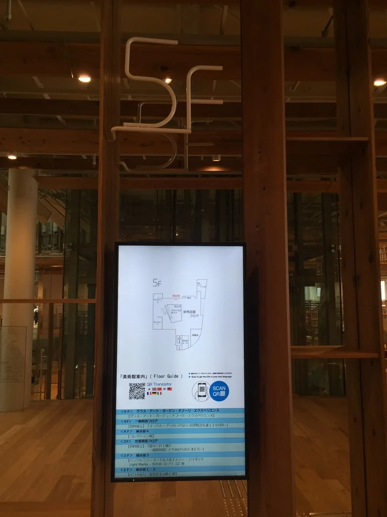 五樓的樓層標示，一根杉木柱上掛著以白色細線寫成的「5F」與一面直立的數位樓層導覽螢幕，背景是玻璃電梯井與杉木的天花