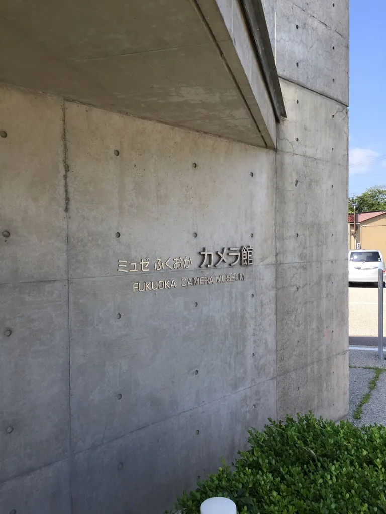 清水混凝土牆面上以金屬字標示館名與英文名，牆面上方是向外出挑的混凝土雨庇，右側是另一道直立的混凝土牆，牆角種著矮灌木，遠處可見停車場與民房
