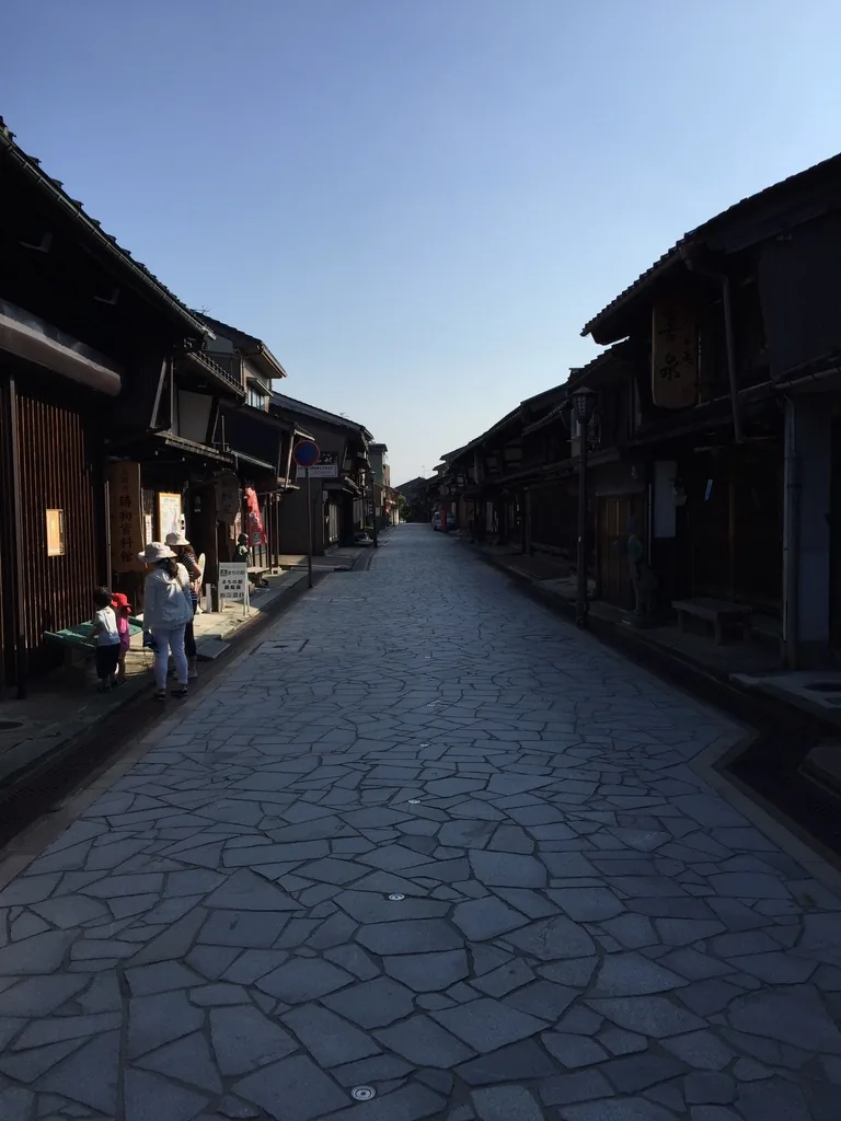 金屋町的石板路，不規則形狀的石材鋪滿整條道路，兩側是木格子立面的兩層町家，屋簷連續，遠處有數名行人