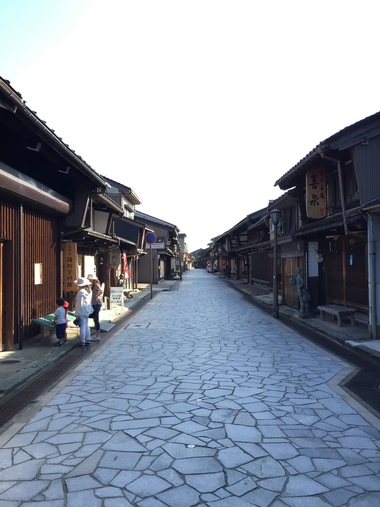 金屋町通的另一個視角，陽光照亮右側的町家與石板路面，左側町家的屋簷下有幾位行人與自行車，路面石材的接縫清晰可見
