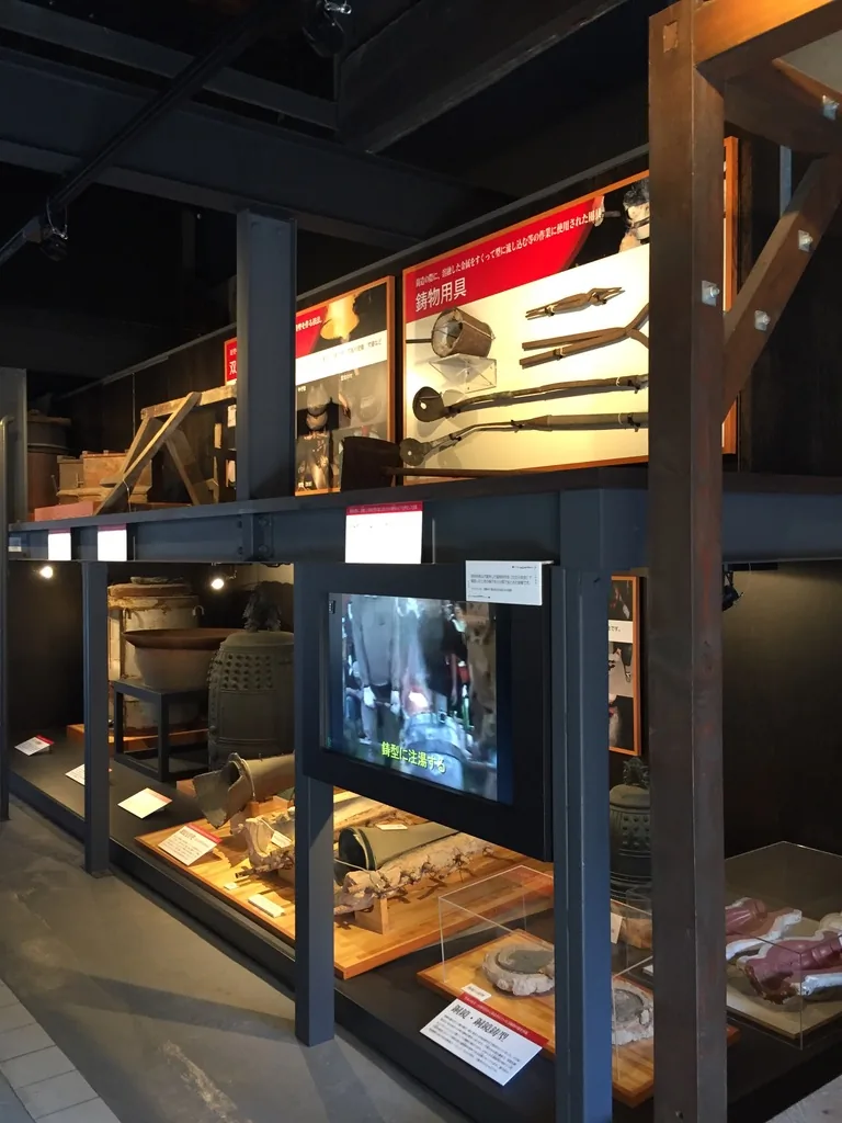 免費展示室的內部，以黑色鋼架組成的展示框分為上下兩層，上層是紅底白字的說明板與鑄造工具，下層是玻璃展示台與播放影片的螢幕，後方可見既存的木柱與斜撐