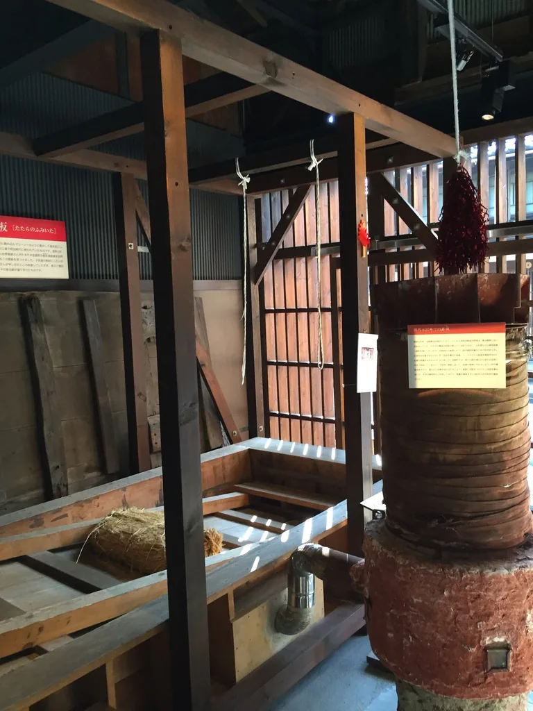 展示室深處，一座以鐵箍箍束的圓筒形爐體立在紅色的磚造基座上，左側是木製的作業台，背景是既存的木柱、斜撐、波浪鐵板牆與木格子窗