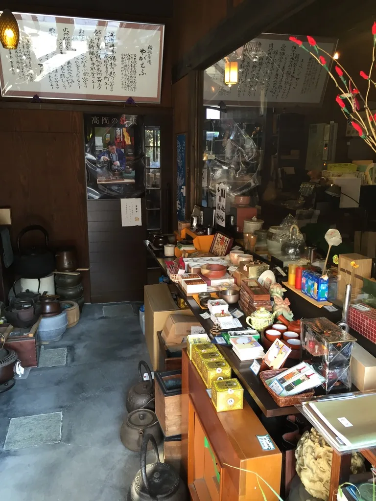 大寺幸八郎商店的店內，土間上陳列著鐵瓶、茶具與銅器小物，牆上掛著裱框的書法，天花是深色的木構件，懸吊著一盞琥珀色的燈具