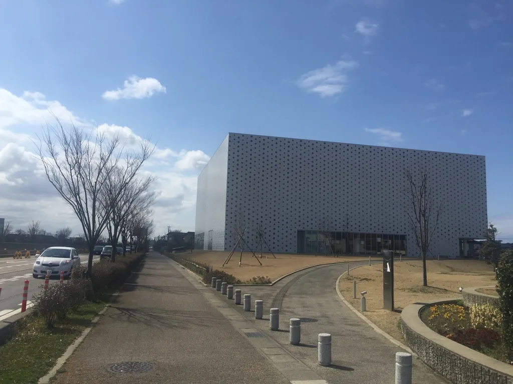 自道路側望向圖書館的引道，左側是行道樹與車道，中央的步道兩側排列著矮石柱，右側一條弧形步道沿草坡繞向入口，遠處是滿布圓孔的白色量體