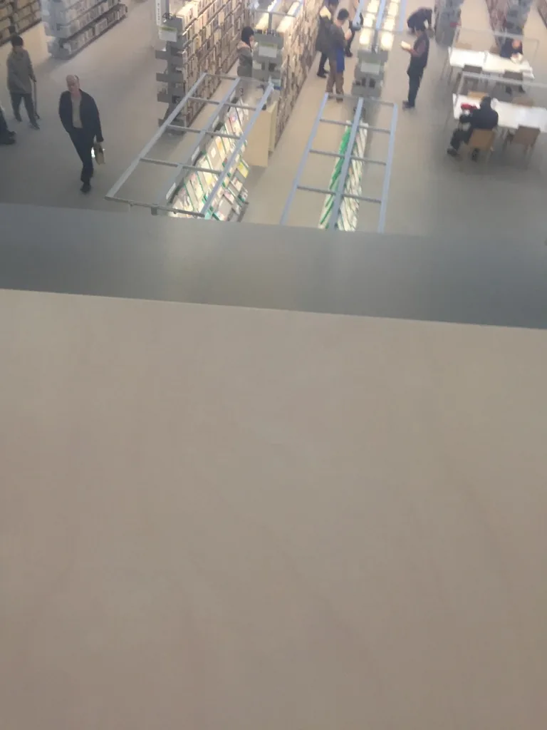 自三樓的扶手邊緣向下俯視，下方是二樓的書架與閱讀桌，幾位讀者在書架間走動，前景是白色的樓板邊緣與玻璃扶手