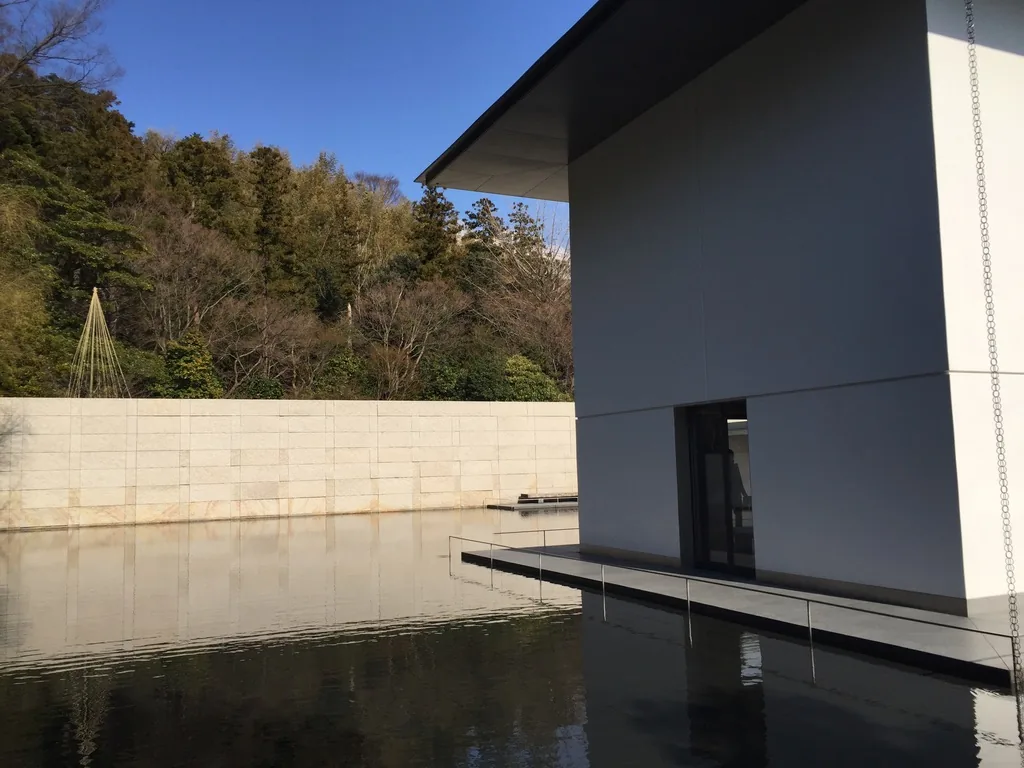 思索空間棟的近景，白色的方形量體立於水面上，上方是向外深出的薄屋簷，量體右側可見一條自簷端垂下的細鍊，前方水面倒映著石牆與天空