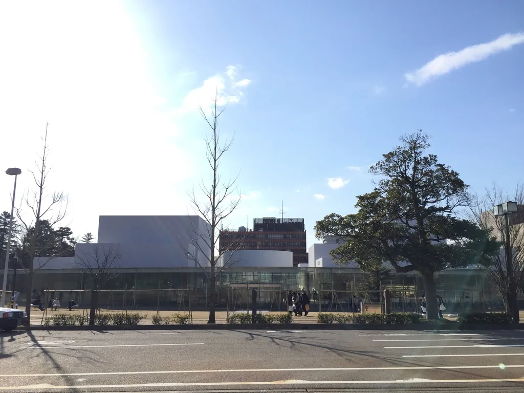 自道路對面看金澤21世紀美術館，低矮的圓形玻璃量體上方突出數個白色方盒，前方是行道樹與寬闊的路面