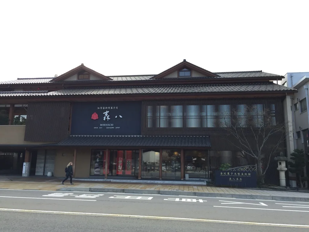 森八本店的正面，三層的和風建築沿道路橫向展開，一樓為玻璃店面與單坡的瓦葺下屋，二樓中央懸掛深色招牌，三樓有兩座小山牆，右側有石燈籠與館名石標