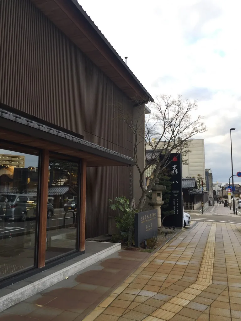 本店側面，二樓的外牆是整面密排的縱向木格柵，一樓為玻璃與木框的店面，上方是瓦葺的單坡屋簷，前方是方磚鋪成的人行道與一座石燈籠