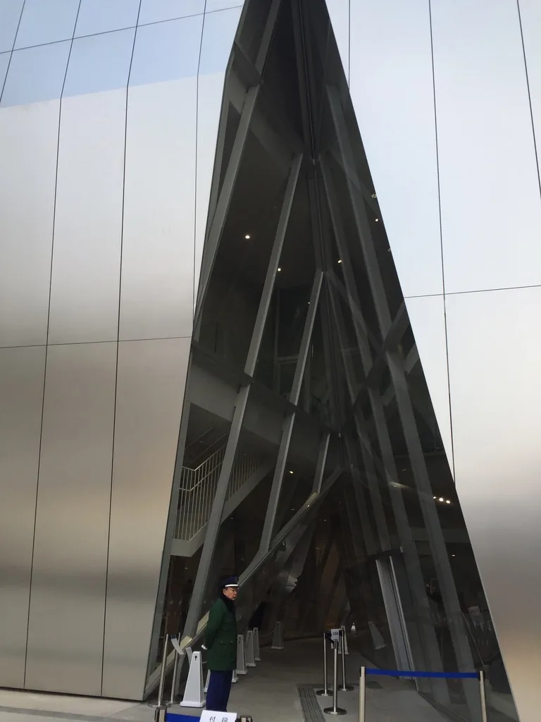 量體之間的縫隙近景，兩片鋁板外牆向內傾斜收成一道三角形的開口，開口內是傾斜的玻璃帷幕與內部的鋼柱，左側可見一位行人