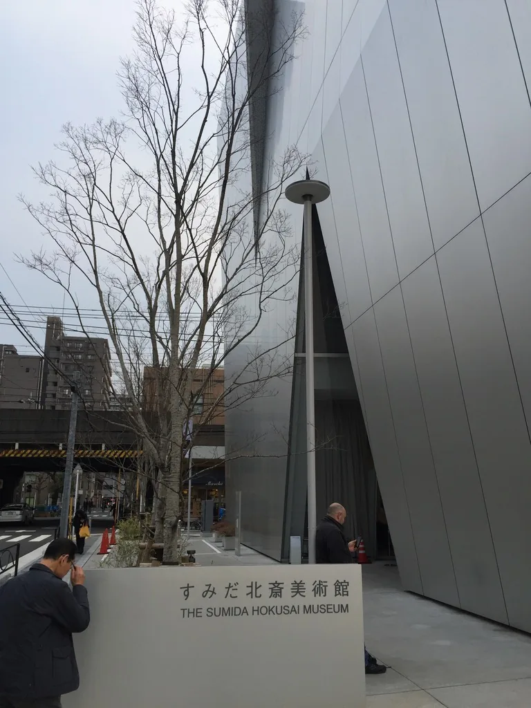美術館一角，鋁板外牆的轉角處切出一道細長的三角形縫隙，前方是一棵落葉的行道樹與一座寫有館名的石材館名台，台前有行人