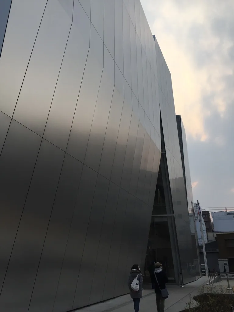鋁板外牆的近景，由下向上仰望，一片片縱向分割的鋁板構成略微傾斜的牆面，牆面上映著模糊的天光與雲，下方有行人
