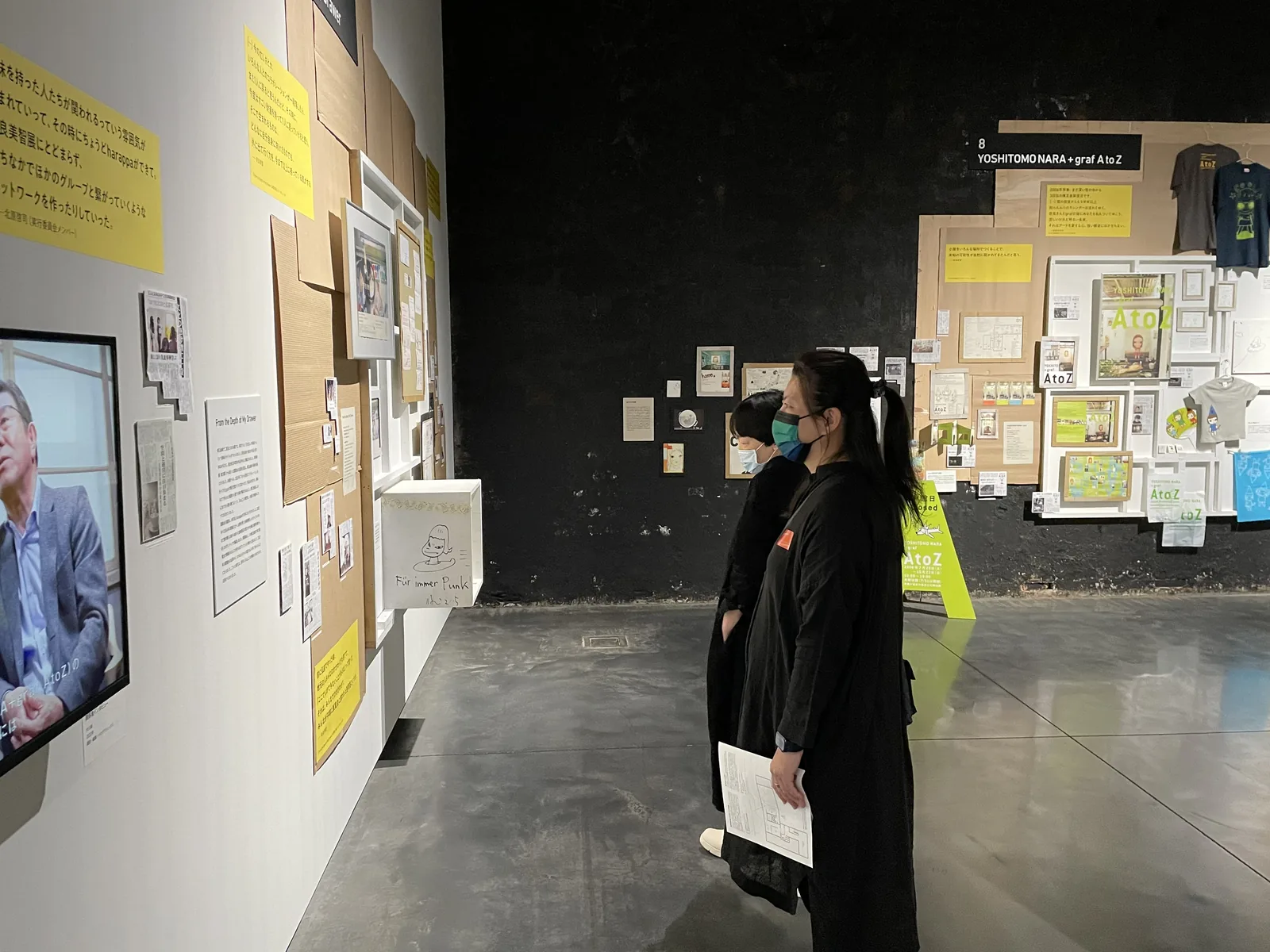 白牆展示室，牆面密排裱框作品與黃色說明卡，地面為深灰色，參觀者沿牆觀看